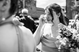 Emotional bride wipes tear from eye in black and white photo outside wedding ceremony.
