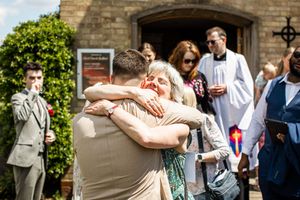 Wedding guests share warm hugs outside stone church entrance with vicar in white robes nearby.