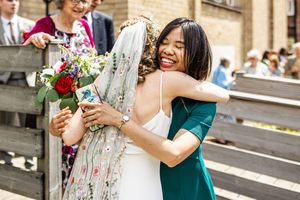 Bride in embroidered veil shares joyful hug with smiling guest in green dress after ceremony.