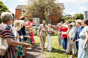 Bride and groom walking through cheering wedding guests outside a red brick house in sunshine.