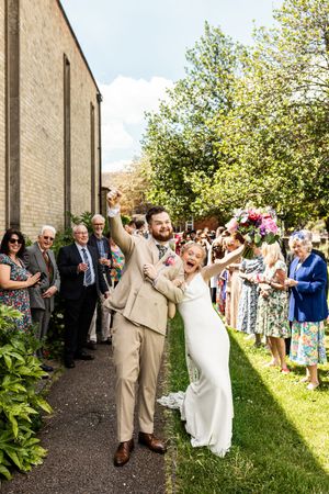 Excited bride and groom celebrate with arms raised walking through confetti line of guests outside church.