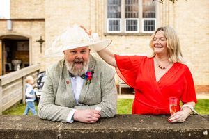 Elegant wedding guest couple, woman in red dress adjusting mans hat outside stone building with champagne.