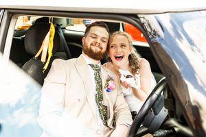Happy newlywed couple sitting in decorated car, groom in cream suit and bride holding keys, smiling.