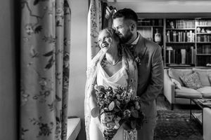 Black and white portrait of bride and groom embracing tenderly by a window with bouquet.