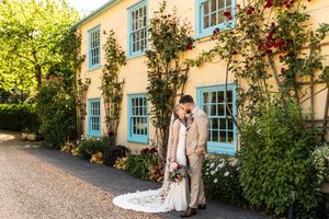 Bride and groom sharing a kiss outside a yellow farmhouse with blue windows and climbing roses.