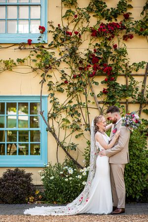 Bride and groom embracing romantically against a yellow wall with red climbing roses and blue windows.
