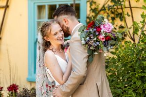 Bride and groom laughing together in a garden beside a yellow building with blue window frames.