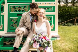 Smiling bride holding colorful bouquet sits with groom on ornate green painted gypsy caravan steps at outdoor wedding.