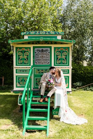 Bride and groom share a kiss seated on steps of decorative green vintage fairground wagon in sunny garden.