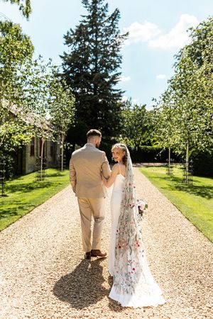 Bride in floral veil and groom walk hand-in-hand down tree-lined gravel path at elegant countryside wedding venue.