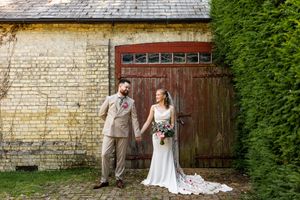 Bride and groom pose before rustic stone barn with wooden doors, bride holding pink and purple bouquet.