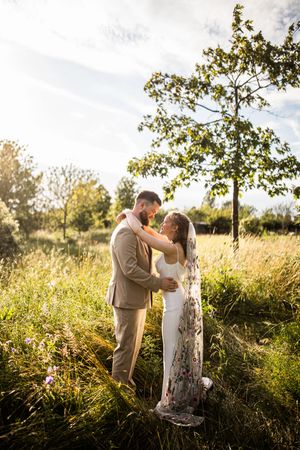 Bride in floral veil and groom embrace in golden sunlit meadow with trees in background at sunset.