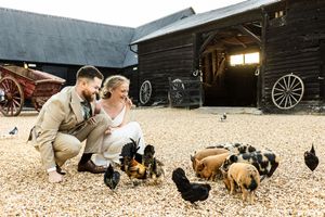 Newlywed couple crouch to feed chickens and piglets outside rustic black barn at countryside farm wedding.