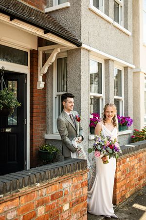 Bride and groom smiling outside a brick house, bride holding a colorful bouquet in a white gown.