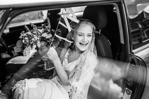 Black and white photo of a laughing bride holding a bouquet while seated in a car on her wedding day.