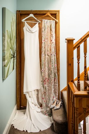 White wedding dress and floral embroidered cape hanging on wooden door frame above bridal heels on stairs.