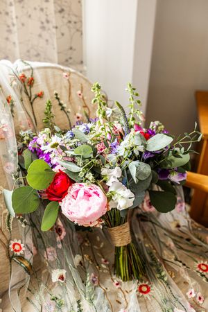 Lush bridal bouquet with pink peonies, purple flowers, and eucalyptus resting on floral embroidered chair.