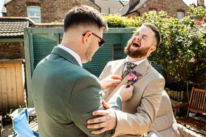 Laughing groom in tan suit getting boutonniere pinned by groomsman in green suit in sunny backyard.