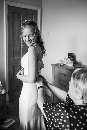 Black and white photo of a smiling bride having her wedding dress fastened by a woman in a polka dot dress.
