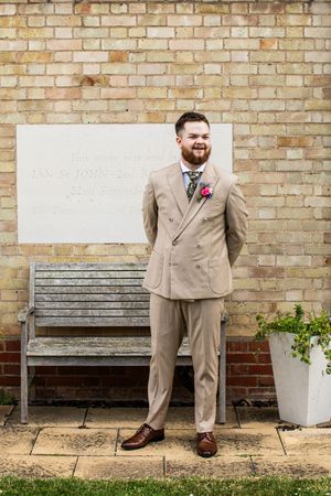 Smiling groom in a beige double-breasted suit with a floral boutonniere standing outside a brick wall.