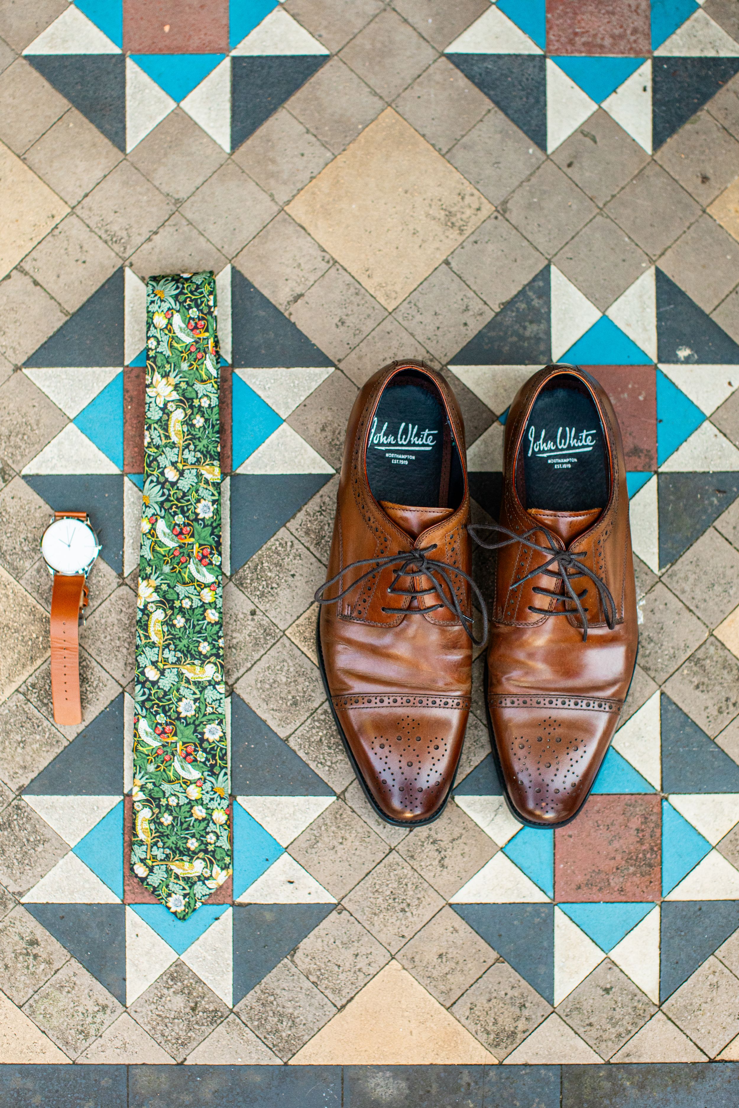 Brown leather brogue shoes, floral tie, and watch arranged on a geometric tiled floor.