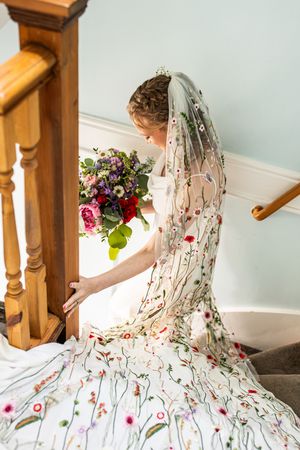 Bride descending a wooden staircase wearing a floral embroidered veil and holding a vibrant bouquet.