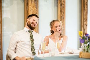 Bride covering mouth laughing while groom smiles during wedding speeches at rustic barn reception.