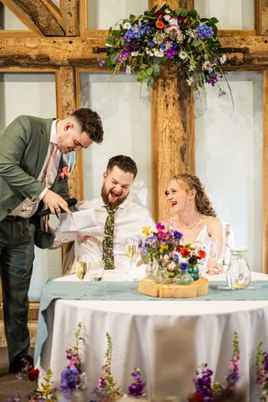 Groom laughing as groomsman pours champagne at the wedding reception table decorated with colorful flowers.