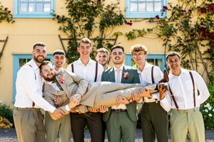 Groom being lifted by groomsmen in suspenders outside a yellow building at a wedding.