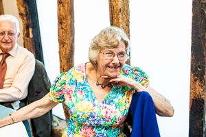 Elderly woman in floral dress laughing joyfully at a wedding reception in a rustic venue.