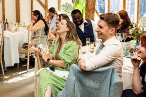 Wedding guests in green dress and suit laughing together during speeches at a bright reception.