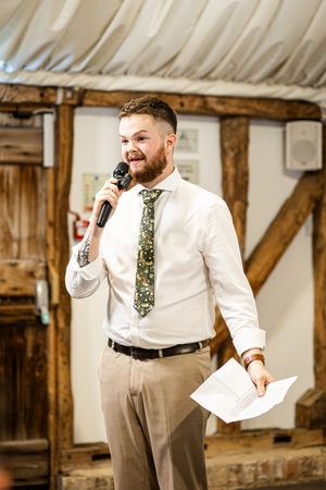 Groom delivering a speech with microphone and notes in hand at a rustic barn wedding reception.