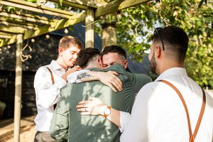 Groomsmen share a heartfelt group hug outdoors under a pergola during wedding celebrations.