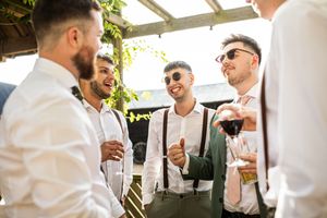 Stylish groomsmen in white shirts and suspenders laughing together outdoors at a wedding reception.