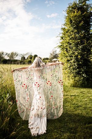 Bride displays stunning floral embroidered wedding veil spread wide in a lush green garden setting.
