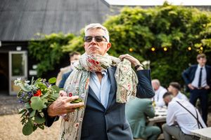 Dapper wedding guest in sunglasses poses confidently with boutonniere and floral scarf outdoors.