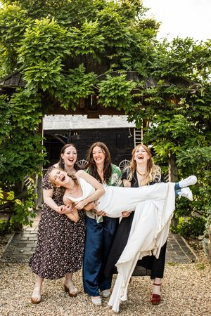 Three friends joyfully lift laughing bride in white dress outside ivy-covered wedding venue.