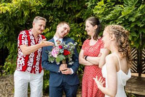 Wedding guests laugh together while admiring bridal bouquet of red and blue flowers outdoors.