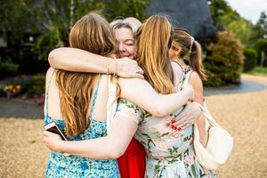 Four women share an emotional group hug on gravel path during outdoor wedding celebration.