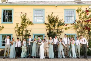 Wedding party in sage green and tan attire posing in front of a yellow venue building.
