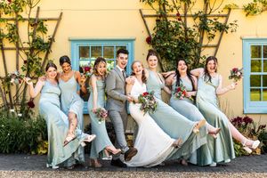 Bride and bridesmaids in sage green dresses laughing and kicking legs up outside yellow venue.