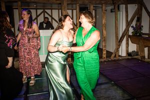Two women dancing together at a wedding reception, one in green jumpsuit and one in silver gown.