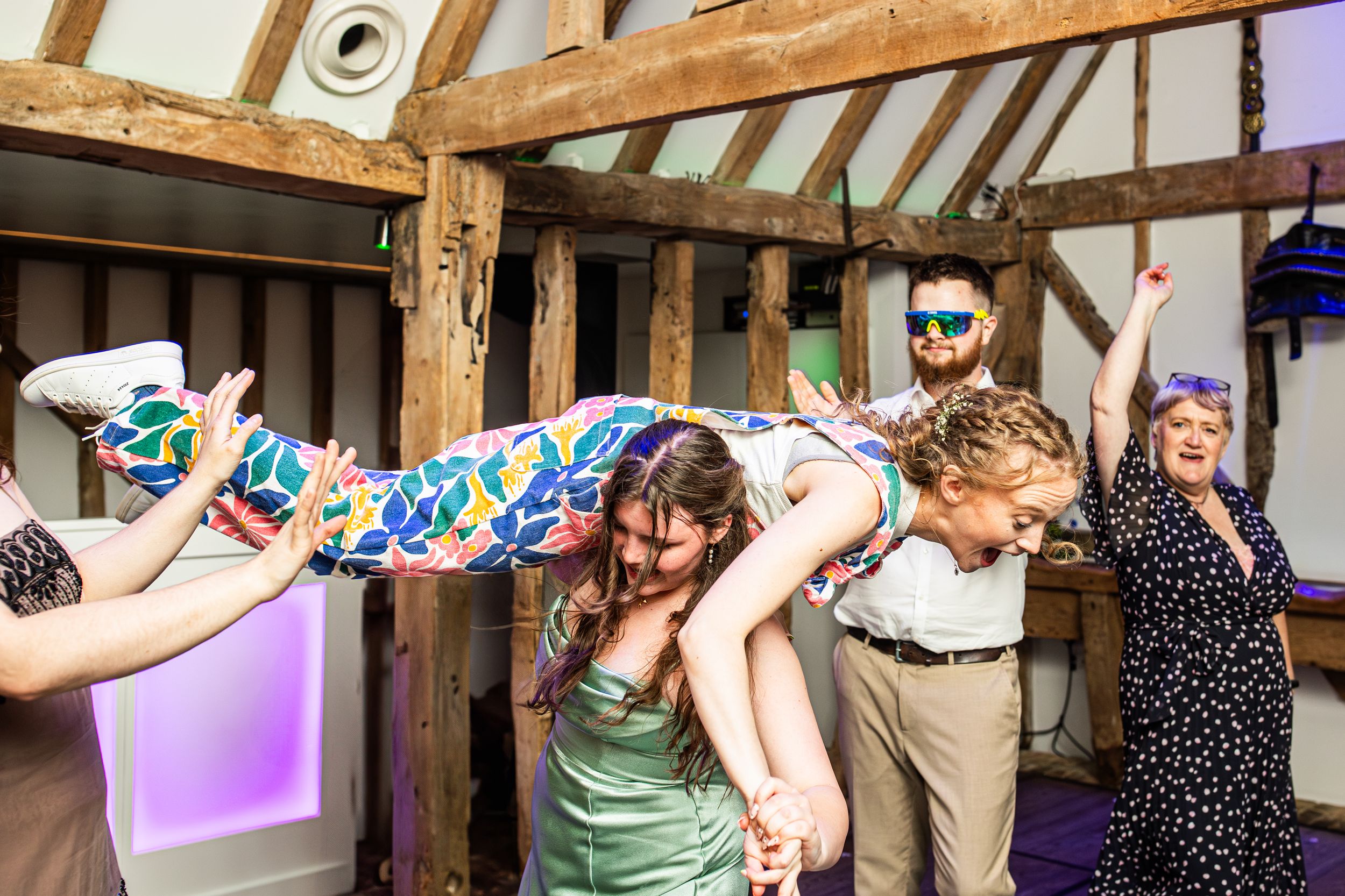 Wedding guests lifting a woman in colorful dress during a lively barn reception celebration.