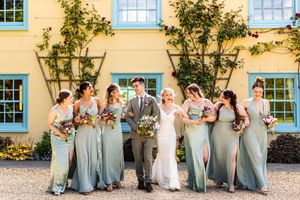 Bride in white gown with groom and six bridesmaids in sage green dresses outside yellow building.