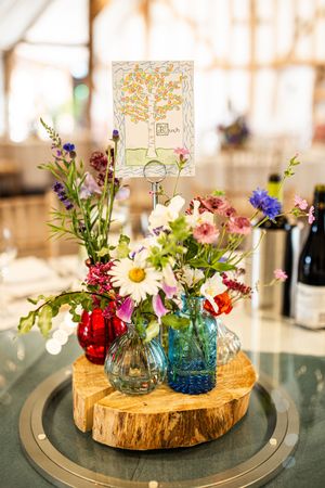 Colorful wildflower centerpiece in mixed glass vases on wood slice with illustrated table number.