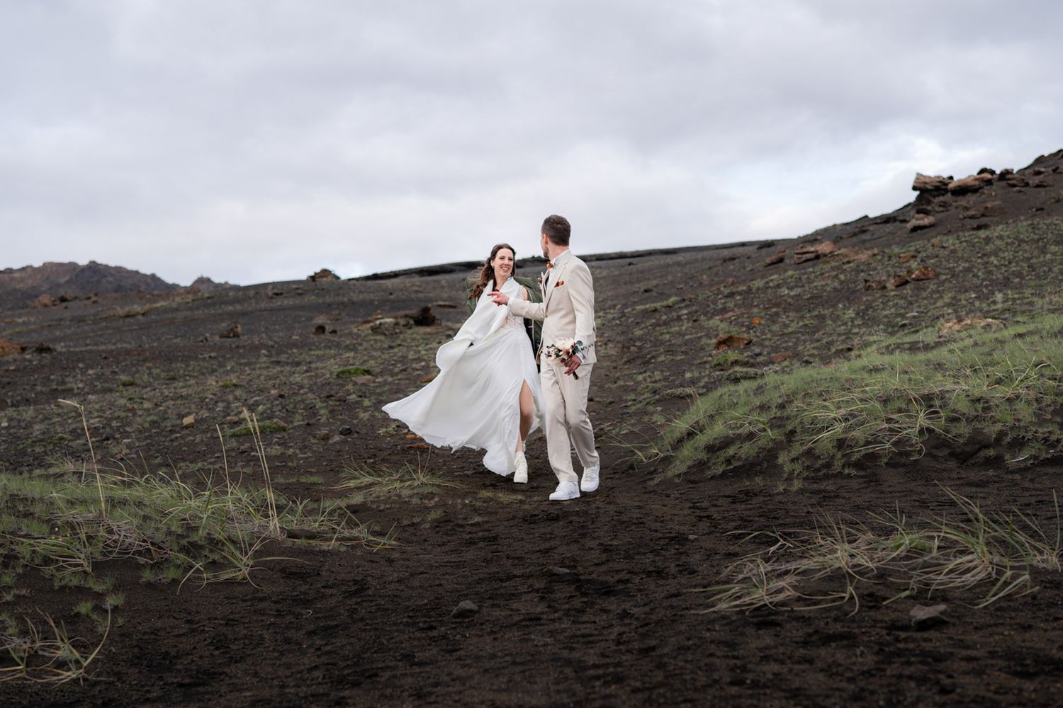 A couple in wedding attire walks along a rugged volcanic landscape with dramatic mountain views in the background.