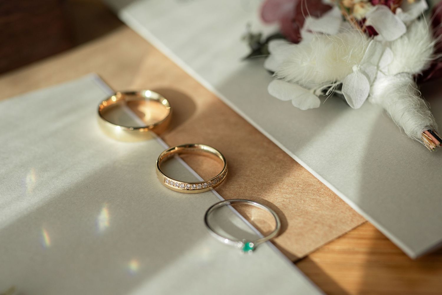Two gold wedding rings rest on a beige surface next to white flower petals in soft natural lighting.