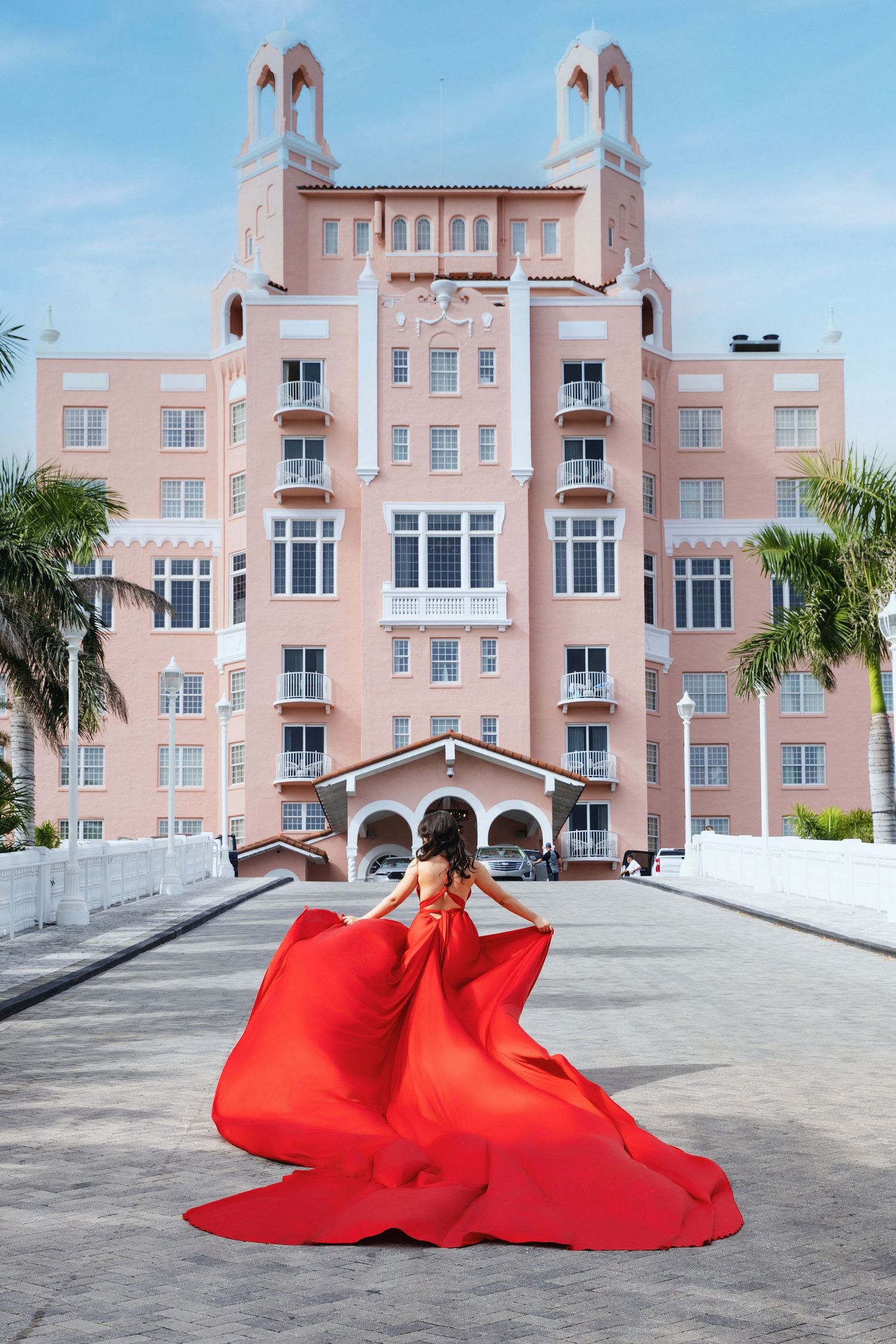 A red fabric flows dramatically across a concrete plaza in front of a pink Art Deco style apartment building with palm trees.