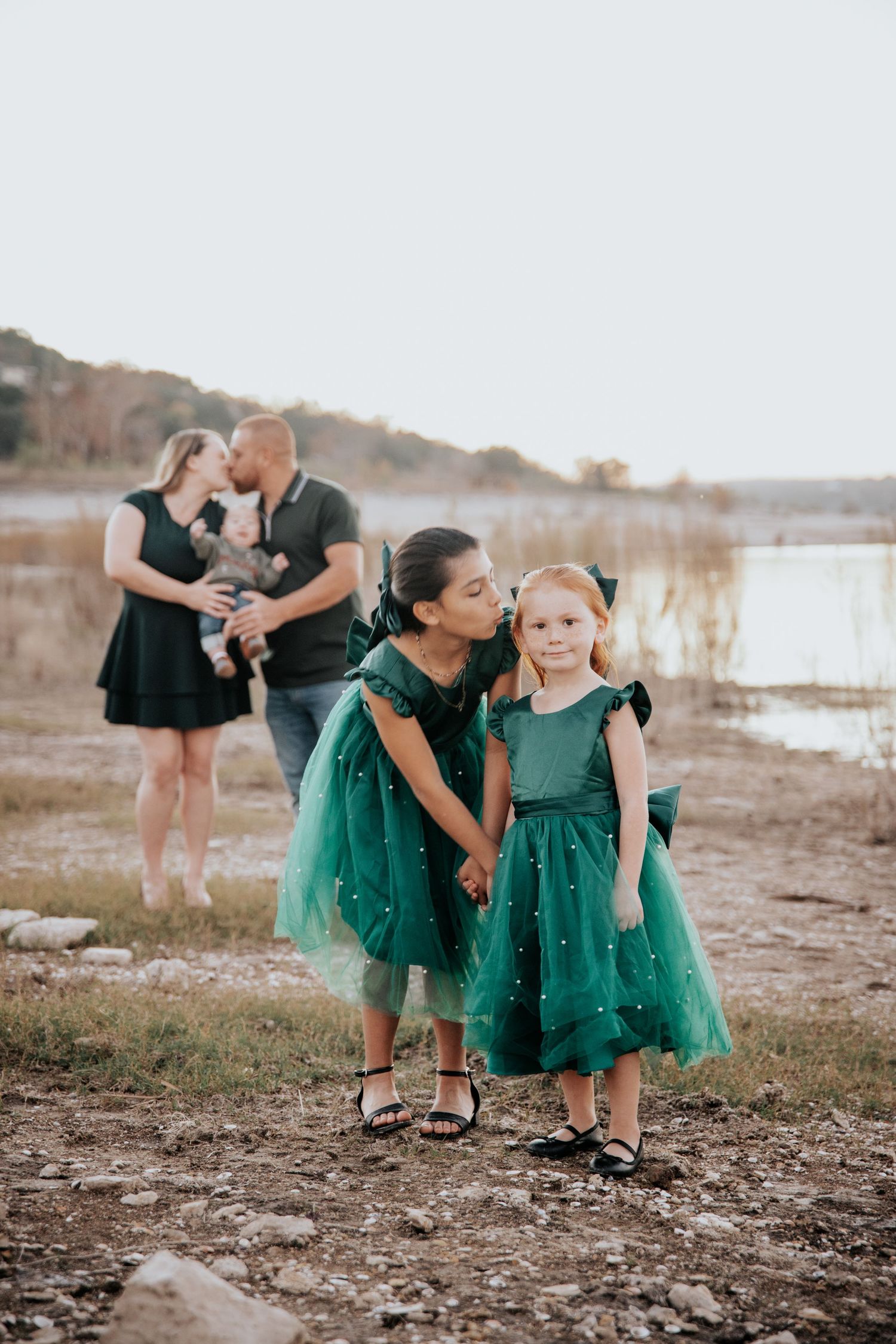 A family in coordinating green outfits poses together during a sunset photoshoot at a lakeside beach.