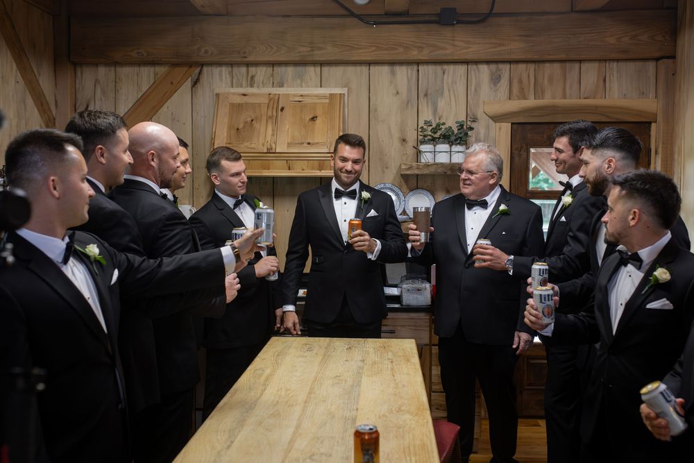 Groomsmen in black tuxedos gather around a wooden table in a rustic venue during pre-wedding celebrations.